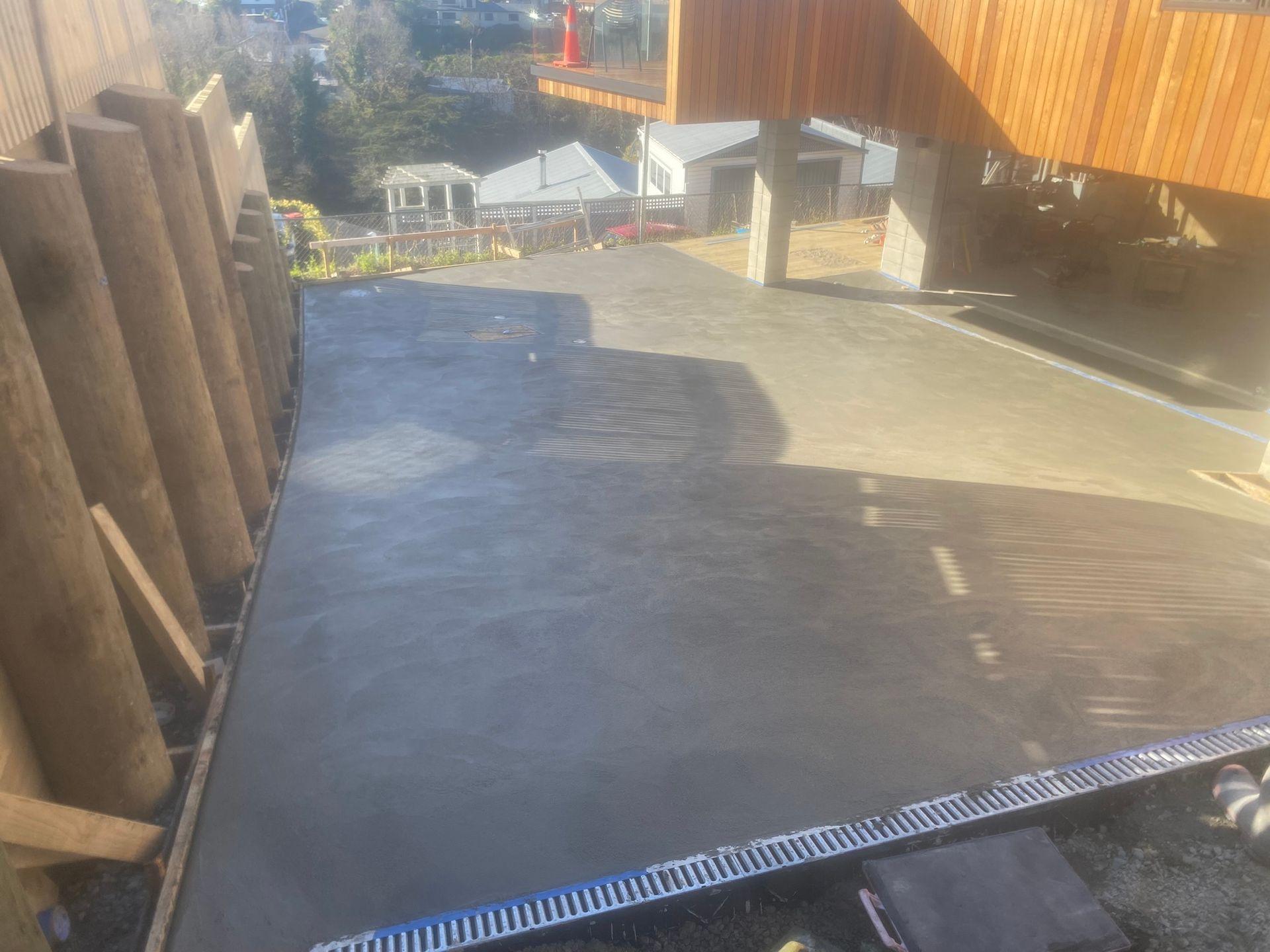 Freshly poured concrete driveway with wooden retaining wall and modern house in background under construction.